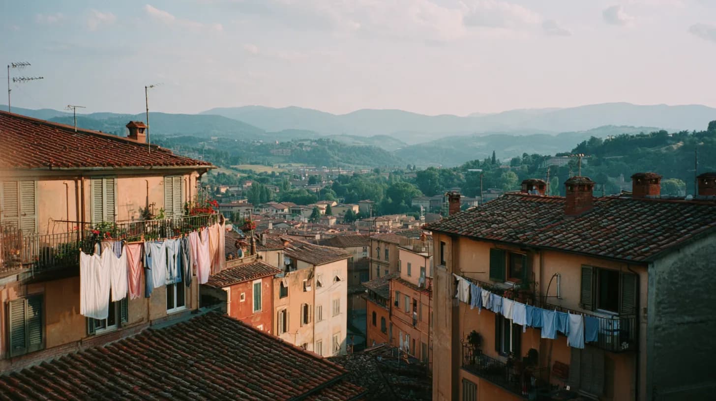 Terracotta rooftops and pastel apartment buildings of a mid-sized Italian city in late afternoon light, with laundry on a balcony and green hills in the distance